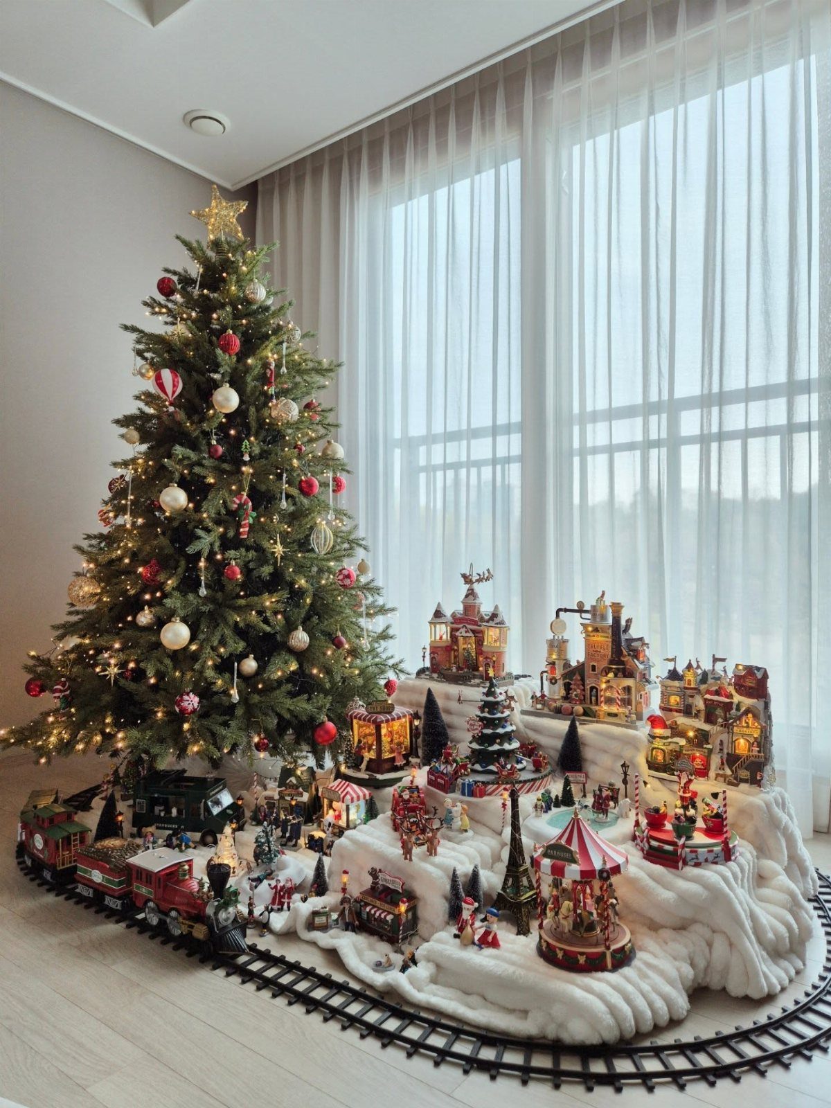 A miniature Christmas village with illuminated houses and a carousel sits beside a decorated tree.