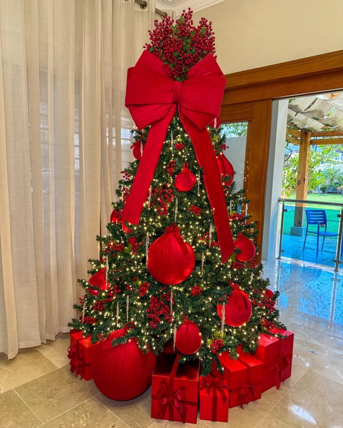 A Christmas tree decorated with red ornaments, a large red bow, and warm lights surrounded by red gifts.