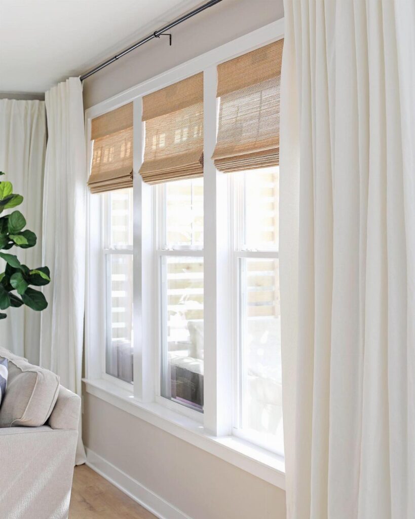 Three tan woven bamboo shades hanging in a white window frame with white curtains.