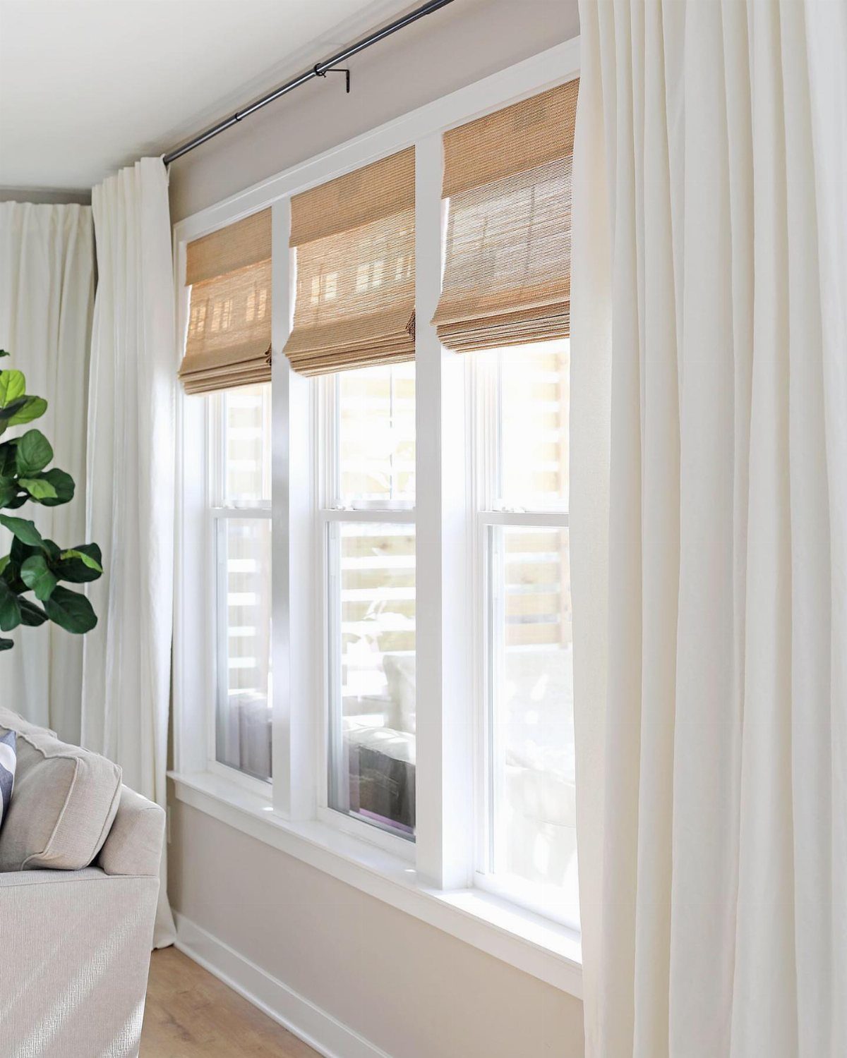 Three tan woven bamboo shades hanging in a white window frame with white curtains.