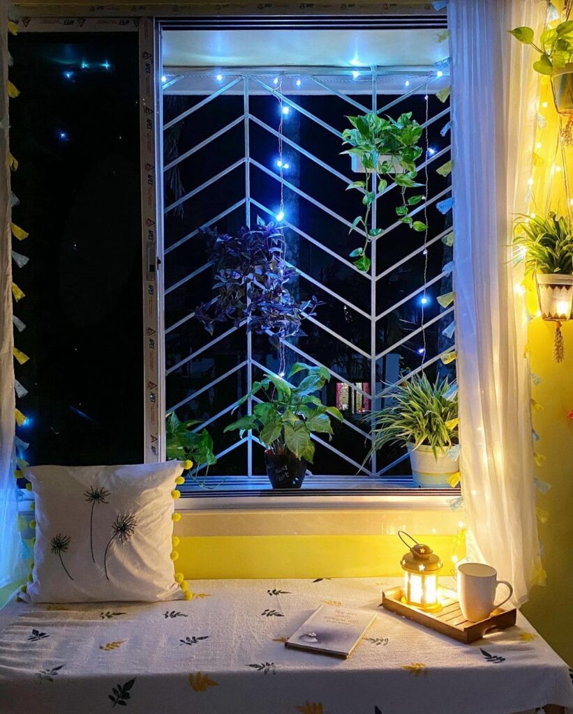 Decorated window with blue fairy lights, hanging plants, and potted greenery on a windowsill at night.