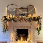 A white fireplace mantel decorated with green garlands, silver ornaments, and glowing white candles.