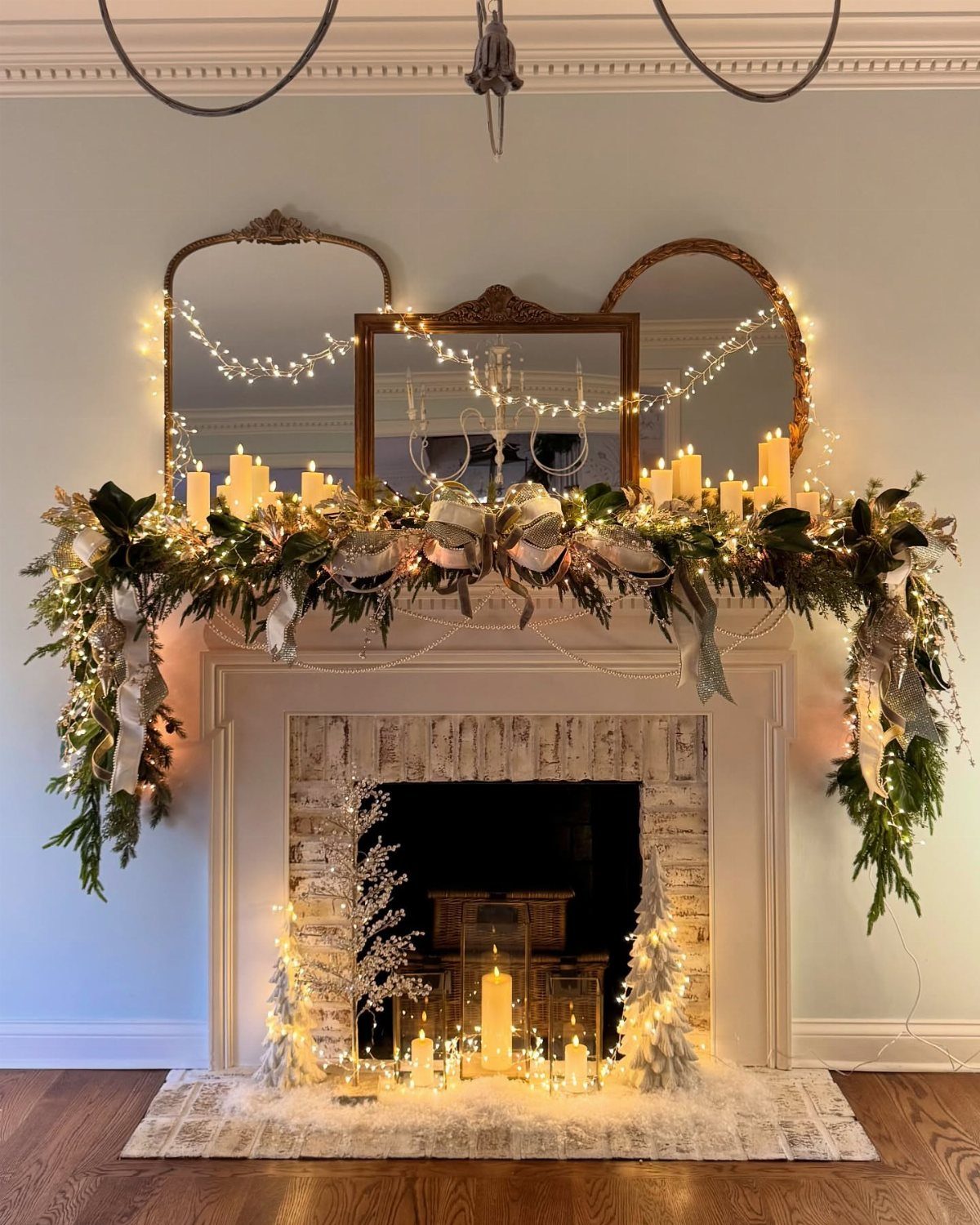 A white fireplace mantel decorated with green garlands, silver ornaments, and glowing white candles.