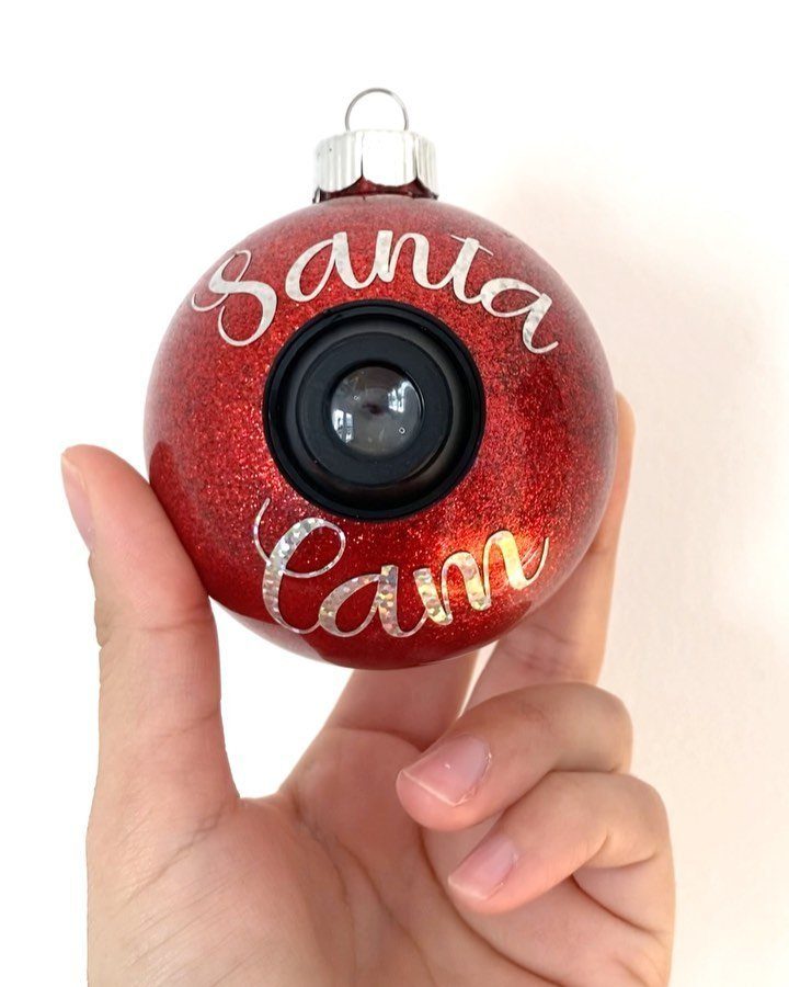 A hand holding a red glittery Santa Cam ornament with a black camera lens.