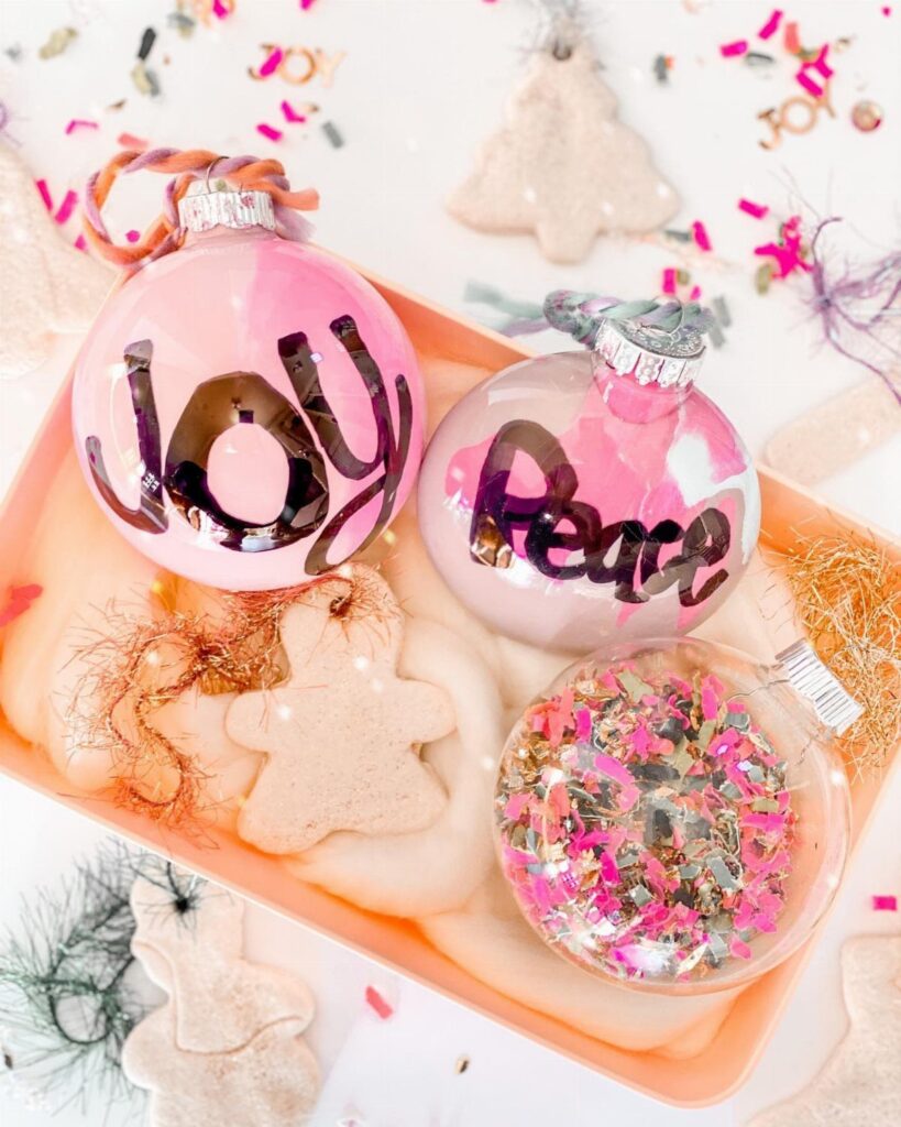 Three decorative glass ornaments resting in a tray, including one filled with pink and gold glitter.