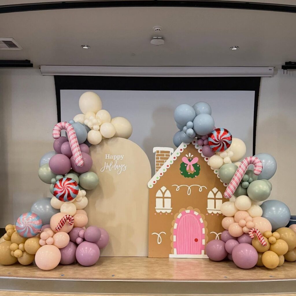 A gingerbread house cutout surrounded by pastel balloon arches and large candy decorations.