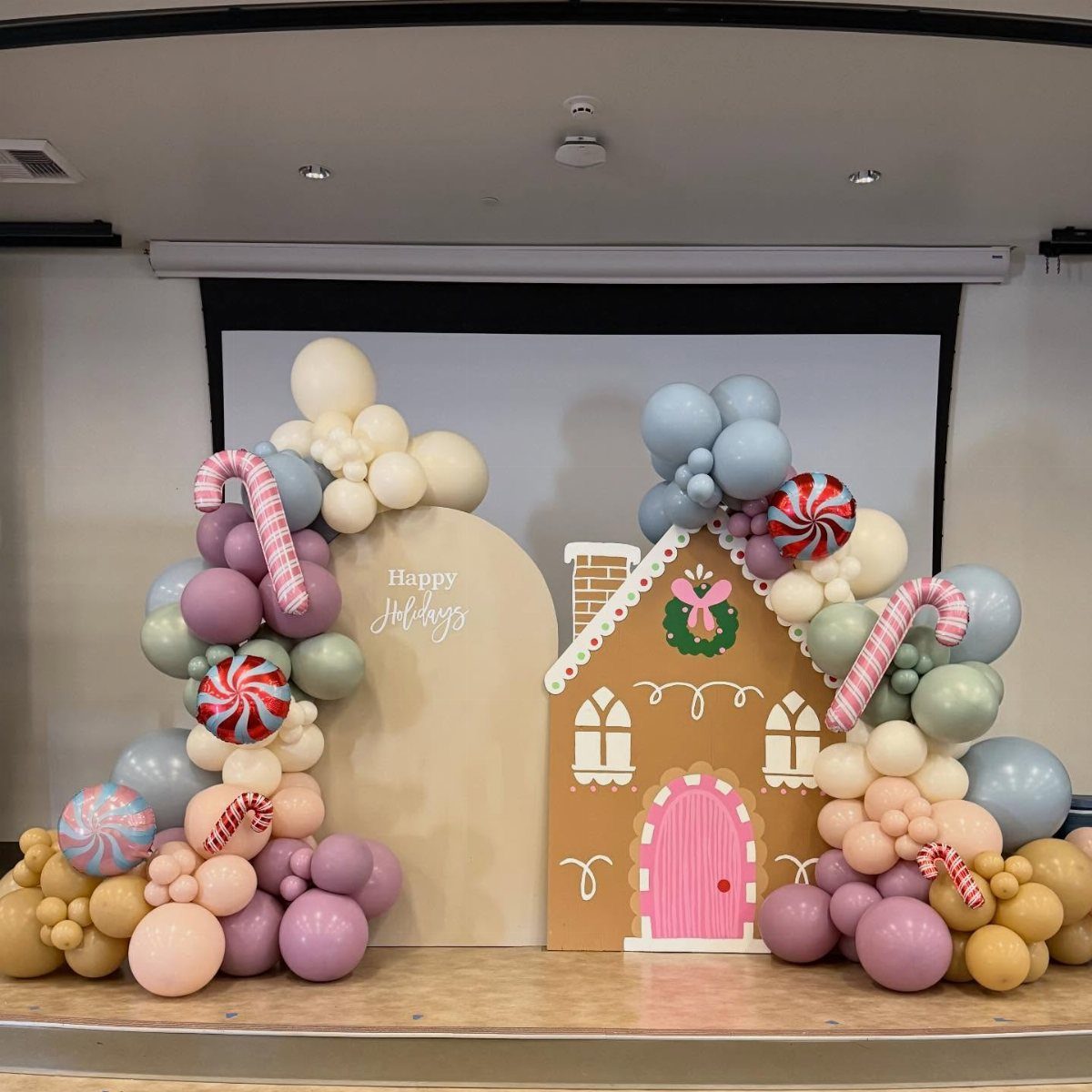 A gingerbread house cutout surrounded by pastel balloon arches and large candy decorations.