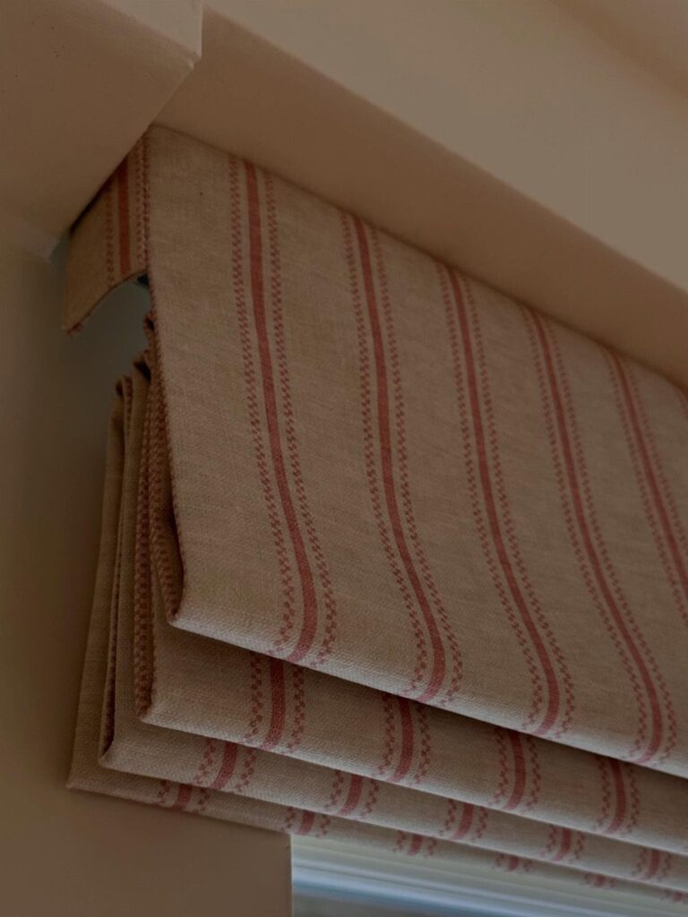 A beige Roman shade with red vertical stripes hanging over a white window frame.