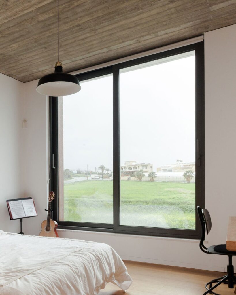 A large black-framed sliding window overlooking a green field from a minimalist bedroom.