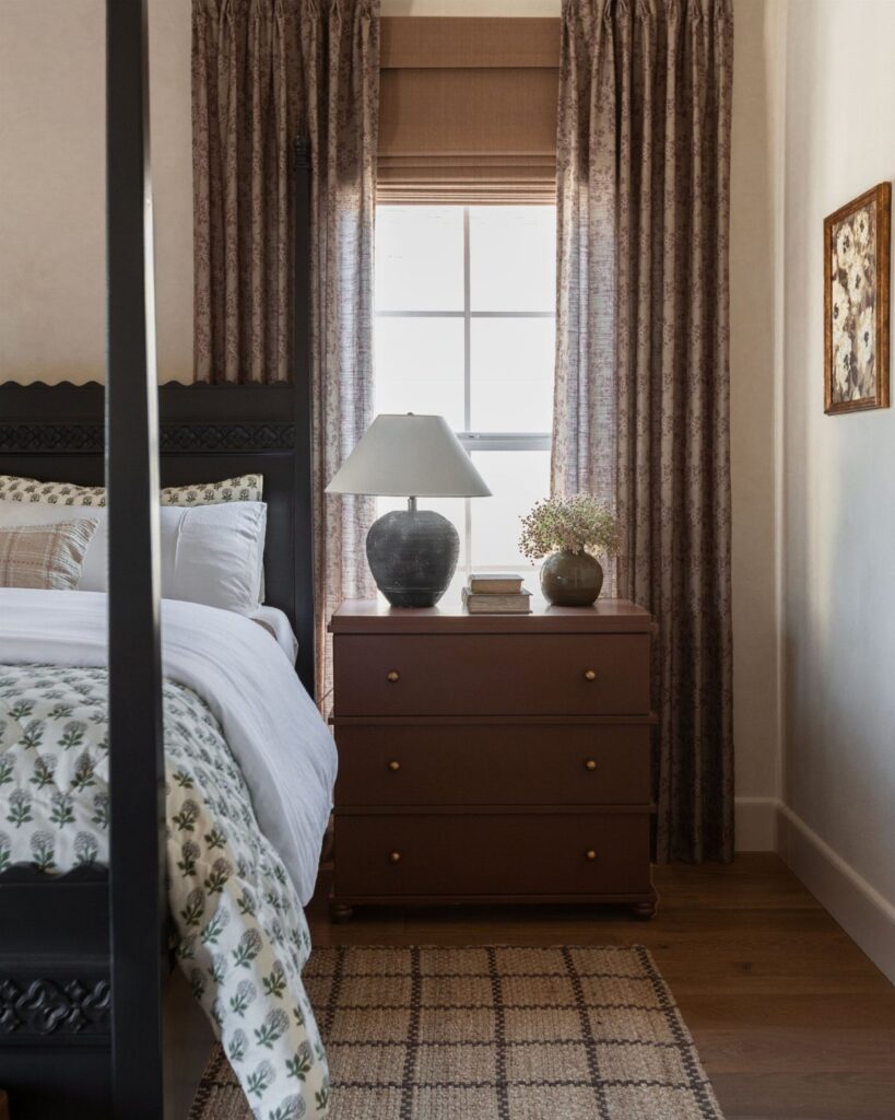 A bedroom window featuring a brown roman shade and long patterned curtains next to a bed.